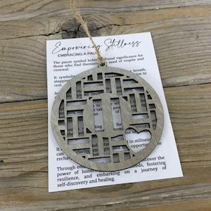 May include: A wooden, gray-toned ornament with a geometric design and a heart-shaped cutout. The ornament hangs from a natural twine string. A white card with the text "Empowering Stillness" and other text is attached.