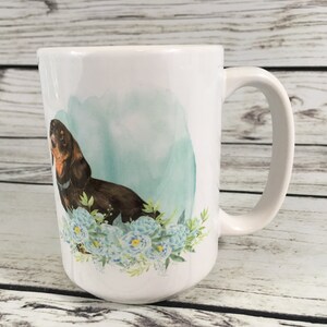 May include: White ceramic coffee mug with a brown and black dachshund dog illustration. The dog is surrounded by blue and green watercolor flowers.
