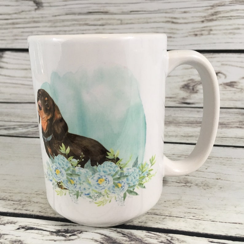 May include: White ceramic coffee mug with a brown and black dachshund dog illustration. The dog is surrounded by blue and green watercolor flowers.