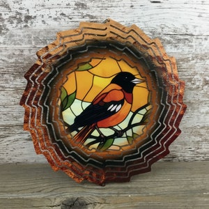 May include: A metal wind spinner with a stained glass design of an orange and black bird perched on a branch. The spinner has multiple layers of metal that create a swirling effect when it spins in the wind.