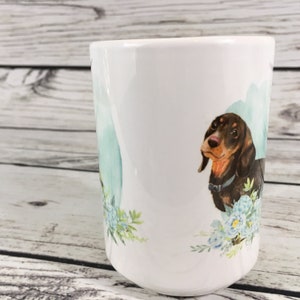 May include: White ceramic mug with a brown and black dachshund illustration. The dachshund is surrounded by blue and green watercolor flowers.