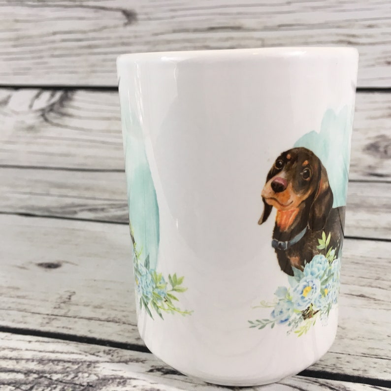 May include: White ceramic mug with a brown and black dachshund illustration. The dachshund is surrounded by blue and green watercolor flowers.