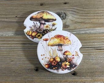 May include: Two round white ceramic coasters with a mushroom and flower design. The coasters feature a yellow and orange mushroom with white spots and a brown stem. The mushrooms are surrounded by yellow and orange flowers and green leaves.