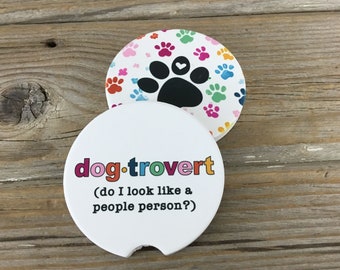 Puede incluir: Un posavasos de cerámica blanco con el texto "dog.trovert (do I look like a people person?)" en colores arcoíris. La parte posterior del posavasos tiene una huella de pata negra con un pequeño corazón blanco sobre un fondo blanco con huellas de pata de colores.