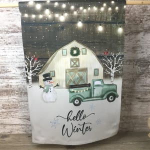 May include: A winter-themed garden flag featuring a barn, snowman, and green pickup truck. The flag has the words "hello Winter" in script. The barn has a wreath on the door, and string lights are above the barn.