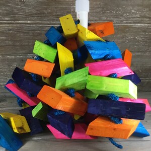 May include: A colorful wooden bird toy with brightly colored blocks connected by blue rope. The blocks are in various shapes and sizes, including rectangles, squares, and triangles. The toy is designed for birds to chew and play with.