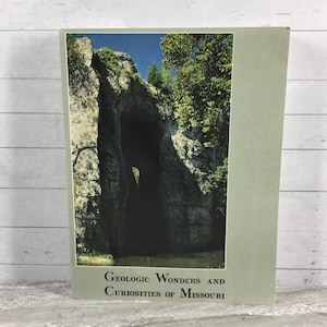 May include: A vintage book titled "Geologic Wonders and Curiosities of Missouri" featuring a landscape image of rock formations and trees. The book has a light green cover and is set against a white wooden background.