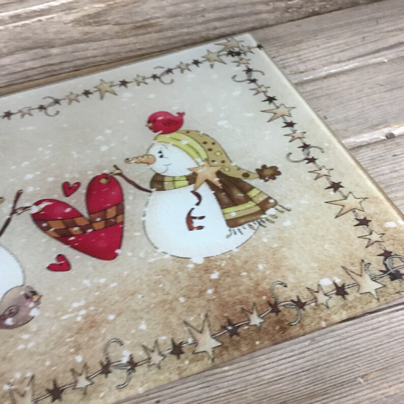 K&ouml;nnte beinhalten: Ein Schneidebrett aus Glas mit Wintermotiv, auf dem zwei Schneem&auml;nner ein rotes Herz halten. Der Hintergrund ist eine braune und wei&szlig;e Schneelandschaft mit einer Sternen-Girlande.