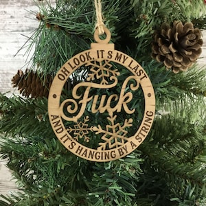 May include: Wooden Christmas ornament with the word "Fuck" in the center, surrounded by the text "Oh look, it's my last and it's hanging by a string." The ornament has snowflake cutouts and hangs from a twine string.