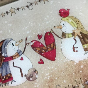 K&ouml;nnte beinhalten: Zwei Schneem&auml;nner halten ein rotes Herz mit einem karierten Muster. Die Schneem&auml;nner tragen Schals und H&uuml;te. Ein Schneemann hat einen roten Vogel auf dem Kopf und der andere einen braunen Vogel zu seinen F&uuml;&szlig;en. Der Hintergrund ist hellbraun mit wei&szlig;en Schneeflocken.