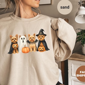 May include: Sand-colored sweatshirt featuring a Halloween-themed graphic. The design includes four Yorkshire Terriers dressed as a pumpkin-carrying dog, a ghost, a bat, and a witch. The sweatshirt is a neutral color, suitable for fall.