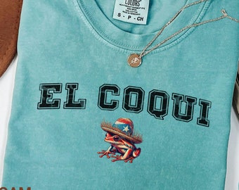 May include: Seafoam green t-shirt with a black and white graphic of a frog wearing a hat with the text "EL COQUI" in black.