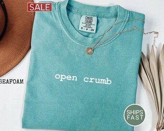 May include: Seafoam green Comfort Colors t-shirt with the words "open crumb" in white. A gold necklace with a pendant rests on the shirt. A "SALE" tag is in the upper left corner. The shirt is folded, and a brown hat is visible.