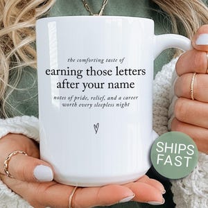 May include: A white ceramic mug featuring black printed text that reads: the comforting taste of earning those letters after your name, notes of pride, relief, and a career worth every sleepless night, with a small hand-drawn heart icon below.