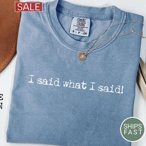 May include: A blue jean colored t-shirt with the phrase "I said what I said!" printed in white. A gold necklace with a pendant rests on the shirt. The shirt has a Comfort Colors tag and a "Ships Fast" sticker.