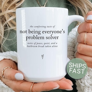 May include: A white ceramic mug featuring black text that reads: the comforting taste of not being everyone's problem solver, notes of peace, quiet, and a bathroom break taken alone, with a small hand-drawn heart symbol below the text.