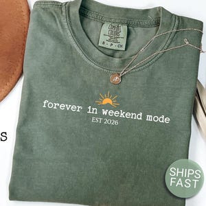 May include: A moss green short-sleeve t-shirt featuring a graphic of a yellow sun above the white text "forever in weekend mode" and "EST 2026". The shirt has a crew neckline and a Comfort Colors brand label printed on the inside of the collar.