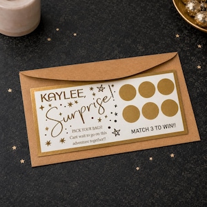 Peut inclure: Une carte à gratter rectangulaire dans une enveloppe en papier kraft. La carte blanche présente un texte en feuille d'or indiquant KAYLEE, Surprise! PACK YOUR BAGS! Cant wait to go on this adventure together!! et MATCH 3 TO WIN!! avec six cercles dorés.
