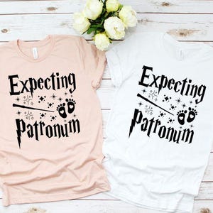 May include: Two t-shirts with the text "Expecting Patronum" in black lettering. The shirts are pink and white and feature a wand, stars, and baby footprints.