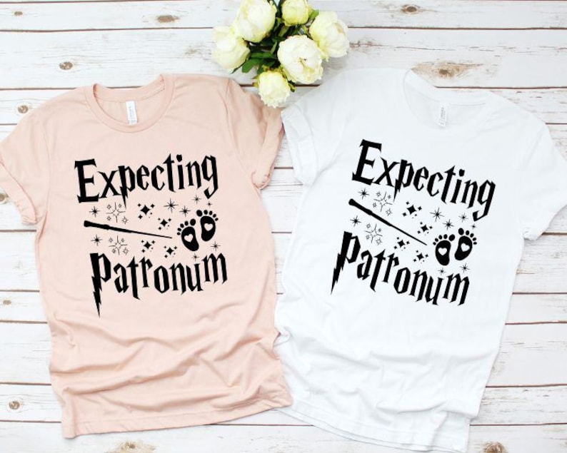May include: Two t-shirts with the text "Expecting Patronum" in black lettering. The shirts are pink and white and feature a wand, stars, and baby footprints.