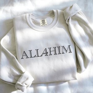 May include: A cream-colored crewneck sweatshirt featuring the black text ALLAHIM printed across the chest. The garment has long sleeves, a ribbed collar, and a Comfort Colors brand label visible on the inside of the neckline showing size M-M.