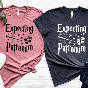 May include: Two t-shirts with the text "Expecting Patronum" and a wand and baby footprints graphic. One shirt is pink and the other is dark blue.