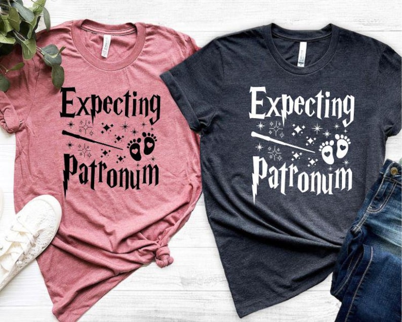 May include: Two t-shirts with the text "Expecting Patronum" and a wand and baby footprints graphic. One shirt is pink and the other is dark blue.