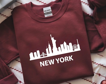 May include: A maroon sweatshirt featuring a white graphic of the New York City skyline, including the Statue of Liberty and the text "NEW YORK". The sweatshirt is made of a soft material.