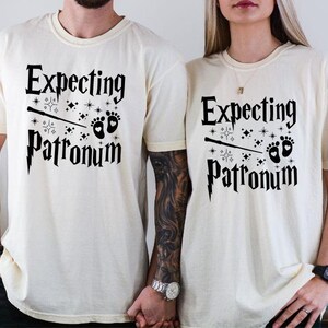 May include: Two white t-shirts with black text that reads "Expecting Patronum" and a graphic of a wand and baby footprints.