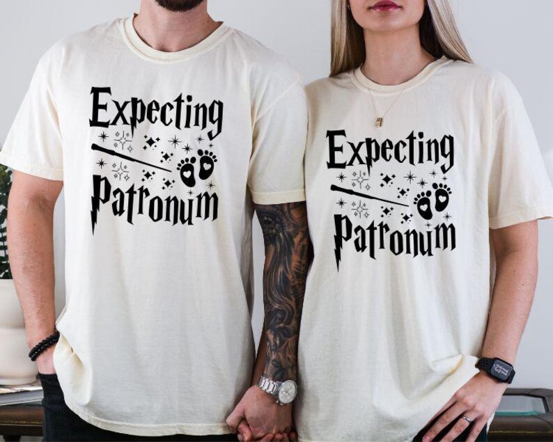 May include: Two white t-shirts with black text that reads "Expecting Patronum" and a graphic of a wand and baby footprints.