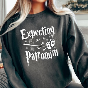 May include: Dark gray sweatshirt with the words "Expecting Patronum" in white, along with a wand, stars, and baby footprints. The sweatshirt is a comfortable, casual style, perfect for expecting parents.