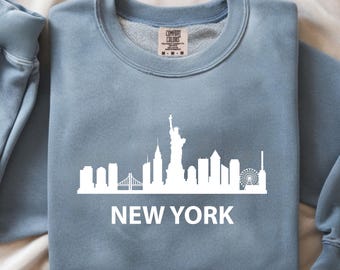 May include: A light blue sweatshirt featuring a white skyline of New York City, including the Statue of Liberty and the text "NEW YORK". The sweatshirt is made of a soft material.