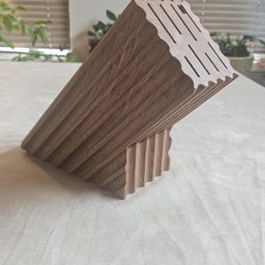 May include: A wooden knife block with a unique, angled design. The block is made of light brown wood and features slots for knives. The design includes a series of vertical grooves and a wavy top edge.