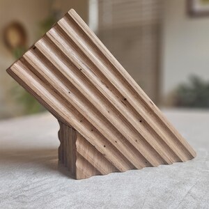 May include: A wooden knife block with a slanted design. The block is made of light brown wood and features a series of parallel grooves on the angled surface. The base has a carved, textured appearance. The knife block is designed to hold knives.