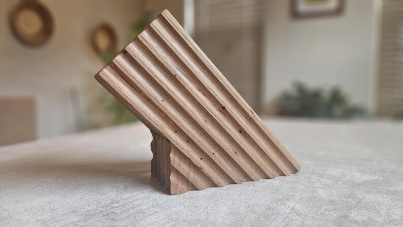 May include: A wooden knife block with a slanted design. The block is made of light brown wood and features a series of parallel grooves on the angled surface. The base has a carved, textured appearance. The knife block is designed to hold knives.