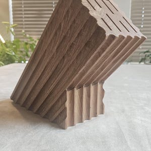 May include: A wooden knife block with a unique, angled design. The block is made of light brown wood and features multiple slots for knife storage. The surface has a grooved texture, adding to its aesthetic appeal. The block is designed to hold various kitchen knives.