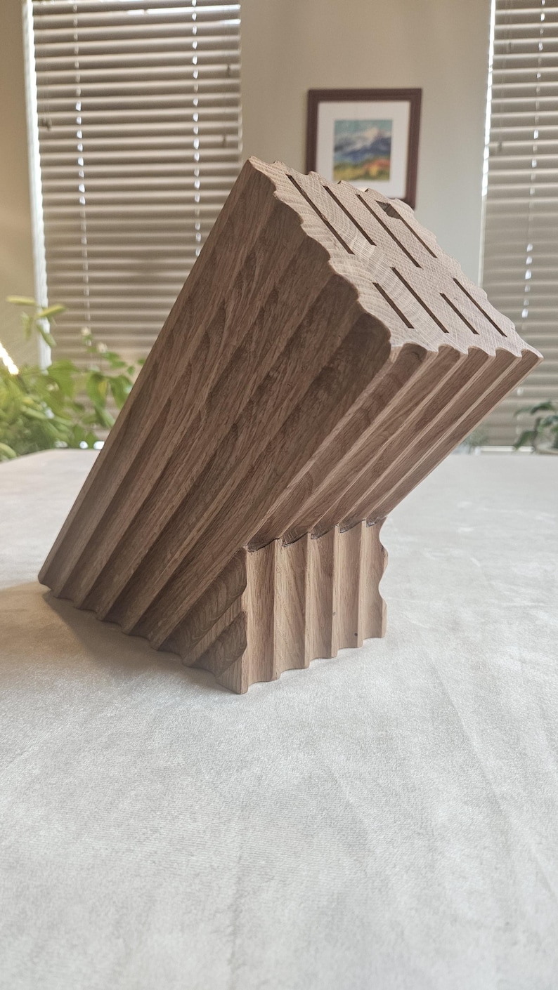 May include: A wooden knife block with a unique, angled design. The block is made of light brown wood and features multiple slots for knife storage. The surface has a grooved texture, adding to its aesthetic appeal. The block is designed to hold various kitchen knives.
