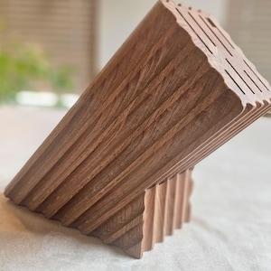 May include: Angled wooden knife block with multiple slots for knife storage. The block is made of a warm-toned wood with a textured, ridged design. The knife block is designed to sit at an angle, showcasing its unique shape and craftsmanship.
