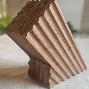 May include: Angled wooden knife block with a natural wood grain finish. The block features multiple slots for knife storage and a unique, sculpted design. The wood has a light brown color and a textured surface, adding to its rustic appeal. The block is designed to hold knives.
