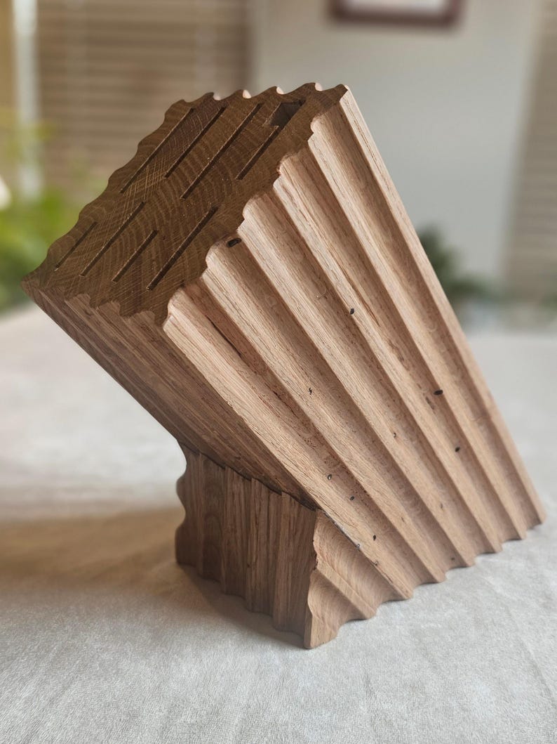 May include: Angled wooden knife block with a natural wood grain finish. The block features multiple slots for knife storage and a unique, sculpted design. The wood has a light brown color and a textured surface, adding to its rustic appeal. The block is designed to hold knives.