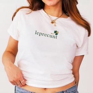 May include: White t-shirt with the word "leprecunt" in a green font, accompanied by a green and gold shamrock design. The shirt is paired with blue denim jeans.