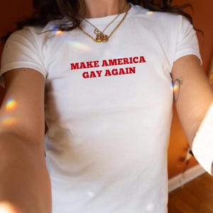May include: White t-shirt with the red text "MAKE AMERICA GAY AGAIN". The shirt has short sleeves and a crew neck. A gold necklace is visible at the neckline.