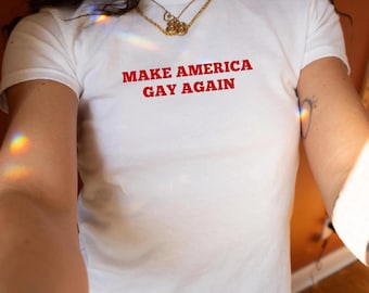 Op de afbeelding: Wit T-shirt met de rode tekst "MAKE AMERICA GAY AGAIN". Het shirt heeft korte mouwen en een ronde hals. Een gouden ketting is zichtbaar bij de halslijn.