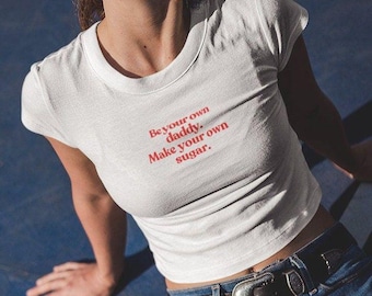 Puede incluir: Top corto blanco con el texto "Be your own daddy. Make your own sugar." en rojo. El top se combina con vaqueros azules y un cinturón negro con hebilla plateada.
