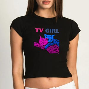 May include: Black cropped t-shirt featuring a graphic of two cats cuddling. The text "TV GIRL" is printed in pink and blue lettering on the shirt.