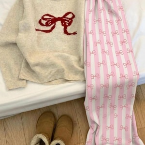 Könnte beinhalten: Ein beiger Pullover mit rotem Schleifendesign, kombiniert mit einer rosa-weiß gestreiften Pyjamahose mit Schleifenmustern. Braune, flauschige Stiefel ergänzen das gemütliche Loungewear-Ensemble. Das Outfit liegt auf einer weißen Oberfläche.