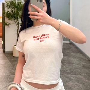 Op de afbeelding: Wit T-shirt met de rode tekst "these titties non-binary too". Het shirt heeft korte mouwen en een ronde hals. De tekst staat in het midden op de borst.