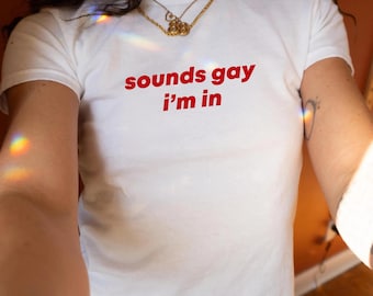 May include: White t-shirt with the red text "sounds gay i'm in". The shirt has short sleeves and a crew neck. A gold necklace is visible at the neckline.
