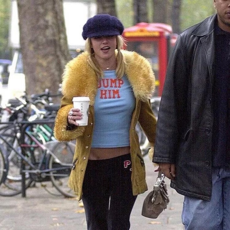 May include: A woman wearing a blue beret, a light blue crop top with the text "JUMP HIM" in red, a tan jacket with a yellow fur collar, and black sweatpants. She is holding a white coffee cup and walking on a city street.