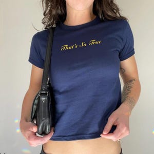Pode incluir: Uma camiseta azul marinho cropped com o texto "That's So True" bordado em dourado na frente.
